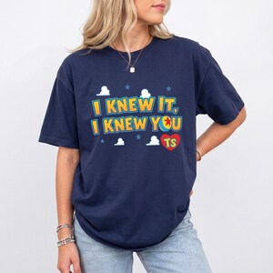K&ouml;nnte beinhalten: Ein dunkelblaues Kurzarm-T-Shirt mit dem gelben Schriftzug "I KNEW IT, I KNEW YOU", einem roten Herz mit den Buchstaben "TS" und einer kleinen Ballgrafik. Das Design enth&auml;lt wei&szlig;e Wolkenformen und kleine blaue Sterne um den Text herum.