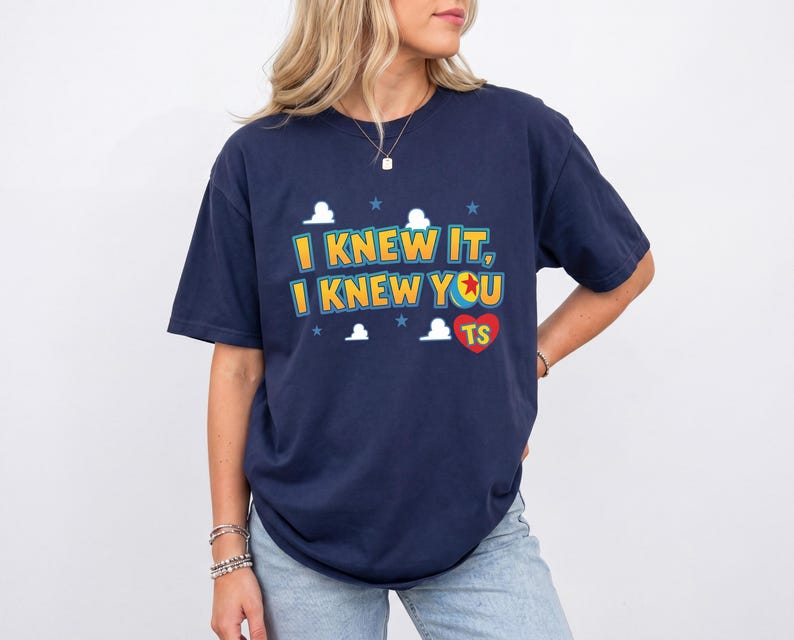 K&ouml;nnte beinhalten: Ein dunkelblaues Kurzarm-T-Shirt mit dem gelben Schriftzug "I KNEW IT, I KNEW YOU", einem roten Herz mit den Buchstaben "TS" und einer kleinen Ballgrafik. Das Design enth&auml;lt wei&szlig;e Wolkenformen und kleine blaue Sterne um den Text herum.