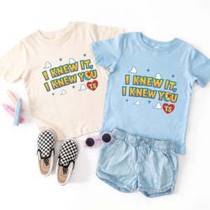 K&ouml;nnte beinhalten: Zwei kurz&auml;rmelige T-Shirts, eines in Creme und eines in Hellblau, jeweils mit dem Text I KNEW IT, I KNEW YOU, einem roten Herz mit den Buchstaben TS sowie kleinen blauen Sternen und wei&szlig;en Wolkenmotiven. Enth&auml;lt zudem karierte Slipper.