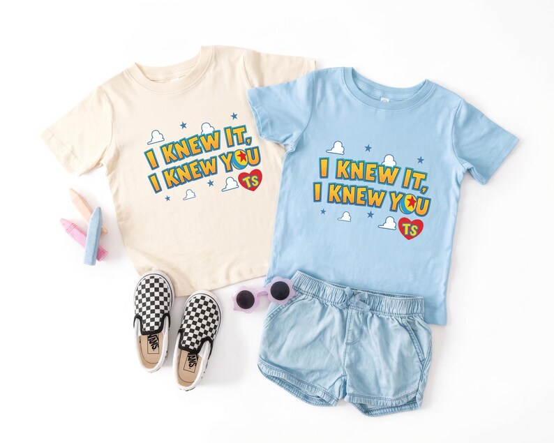 K&ouml;nnte beinhalten: Zwei kurz&auml;rmelige T-Shirts, eines in Creme und eines in Hellblau, jeweils mit dem Text I KNEW IT, I KNEW YOU, einem roten Herz mit den Buchstaben TS sowie kleinen blauen Sternen und wei&szlig;en Wolkenmotiven. Enth&auml;lt zudem karierte Slipper.