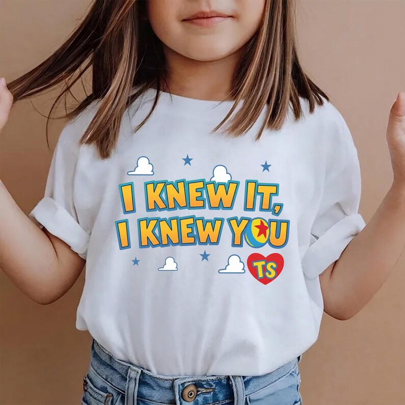 K&ouml;nnte beinhalten: Ein wei&szlig;es T-Shirt mit kurzen &Auml;rmeln und dem gelben Schriftzug I KNEW IT, I KNEW YOU in blauer Umrandung. Das Design auf der Vorderseite zeigt zudem ein rotes Herz mit den Buchstaben TS, kleine blaue Sterne und wei&szlig;e Wolkenformen.