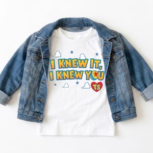 K&ouml;nnte beinhalten: Ein wei&szlig;es kurz&auml;rmeliges T-Shirt mit dem Text I KNEW IT, I KNEW YOU, einer bunten Ballgrafik und einem roten Herz mit den Buchstaben TS, umgeben von blauen Sternen und wei&szlig;en Wolkenumrissen, getragen unter einer blauen Jeansjacke.
