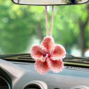 May include: A handmade, pink crocheted flower car charm hanging from a rearview mirror. The flower has five petals and a small, red center. The charm is suspended by a matching pink cord.
