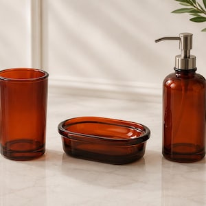 Può includere: Un set da bagno con un bicchiere, un portasapone e un flacone con dosatore. Gli articoli sono in vetro color ambra. Il flacone con dosatore ha una pompa color argento. Il set è su una superficie di marmo.