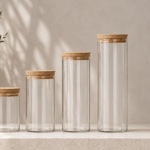 Peut inclure: Quatre bocaux de rangement cylindriques en verre transparent de hauteurs différentes, chacun surmonté d'un couvercle en liège marron clair. Les bocaux sont disposés en rangée du plus petit au plus grand, avec un design simple et transparent.
