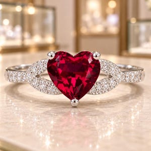 Puede incluir: Un anillo de tono plateado con una gran piedra preciosa roja facetada en forma de corazón como pieza central. La banda está adornada con pequeñas piedras claras engastadas en pavé que se extienden desde los lados del corazón central, creando un diseño simétrico.