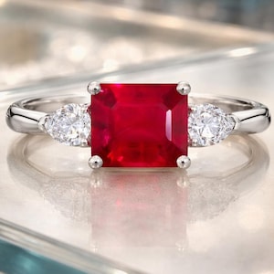 Puede incluir: Un anillo de tono plateado con una gran piedra preciosa roja vibrante de corte cuadrado en el centro, flanqueada por dos piedras blancas transparentes más pequeñas en forma de pera a cada lado. La banda es lisa y pulida con engaste de garras.