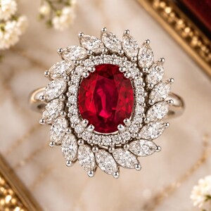 May include: A ring featuring a large oval-cut red gemstone at the centre, surrounded by a double halo of small round diamonds and an outer border of marquise-cut diamonds set in a polished silver-toned metal band with a floral-inspired design.