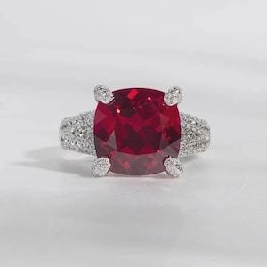 May include: A silver-toned ring featuring a large, cushion-cut, deep red gemstone held by four prongs. The band and the prongs are encrusted with numerous small, clear, sparkling stones that extend along the sides of the ring's metal shank.