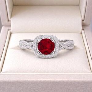 Puede incluir: Un anillo de tono plateado con una piedra central redonda de color rojo intenso, rodeada por un halo de pequeñas piedras claras y brillantes. La banda está dividida y adornada con piedras claras adicionales, creando un aspecto texturizado y brillante.