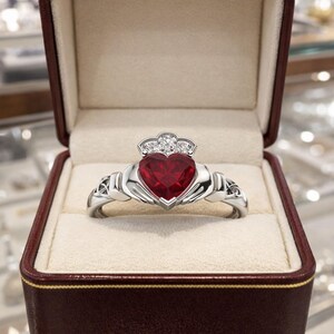 Op de afbeelding: Een zilveren Claddagh-ring met een hartvormige rode edelsteen in het midden, bekroond met een kleine kroon bezet met drie heldere stenen. De ringband heeft gedetailleerde knoopmotieven aan beide zijden en zit in een donkerrood sieradendoosje.