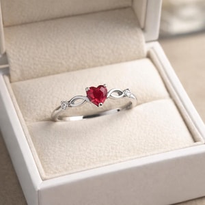 Puede incluir: Un anillo de tono plateado con una piedra preciosa roja central en forma de corazón. La banda tiene un diseño de infinito a ambos lados de la piedra central, acentuado con dos pequeñas piedras redondas transparentes engastadas.