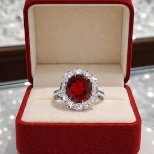 May include: A silver-toned ring featuring a large, oval-cut deep red gemstone at the center, surrounded by a halo of smaller, round, and baguette-cut clear stones. The ring is presented inside an open, rectangular, red velvet jewelry box.