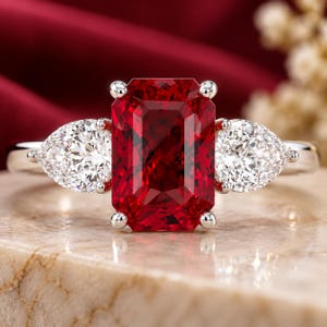 Puede incluir: Un anillo de tono plateado con una gema grande, rectangular y de color rojo intenso en el centro, flanqueada por dos gemas blancas transparentes más pequeñas en forma de pera a cada lado, aseguradas con garras sobre una banda metálica pulida.