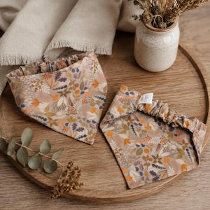 May include: Two triangular pet bandanas featuring a repeating pattern of autumn-coloured leaves, berries, and botanical elements in shades of orange, brown, and muted purple on a light beige background, each with an elasticated neck opening.