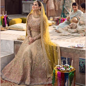 Könnte beinhalten: Ein bodenlanges beiges Kleid mit aufwendigen goldenen und rosafarbenen Stickereien, langen Ärmeln und einem figurbetonten Oberteil. Das Kleid wird mit einem transparenten, leuchtend gelben Dupatta kombiniert, das über Kopf und Schultern liegt.