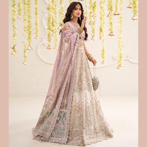 May include: A floor-length lehenga choli set in shades of cream, beige, and soft lavender. The ensemble features intricate silver and gold embroidery, beadwork, and sequins throughout the skirt and bodice, paired with a sheer, embellished dupatta.