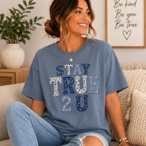 Può includere: Una t-shirt a maniche corte color blu polvere con la scritta STAY TRUE 2 U in stile patchwork. Le lettere sono riempite con vari motivi blu e bianchi, tra cui stampe floreali e quadretti vichy, creando un aspetto strutturato e stratificato.