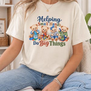 May include: A cream-colored short-sleeve t-shirt featuring a graphic of four cartoon teddy bears engaged in various activities. The text "Helping Small Hands Do Big Things" is printed in colorful lettering above and below the bear illustration.