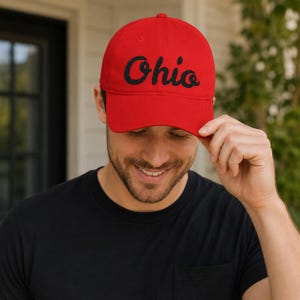 Könnte beinhalten: Eine leuchtend rote Baseballkappe mit dem Wort Ohio in schwarzer Schreibschrift auf der Vorderseite. Die Kappe hat eine strukturierte Krone, einen gebogenen Schirm und einen Knopf oben in der Mitte für einen klassischen Look und Sitz.