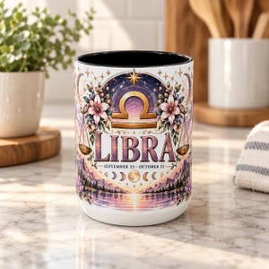 Könnte beinhalten: Ein weißer Keramikbecher mit schwarzem Innenraum und einem Waage-Sternzeichen-Motiv. Die Grafik zeigt das Waage-Symbol, florale Akzente, einen Mondphasenzyklus und den Text "LIBRA SEPTEMBER 23 - OCTOBER 22" über einer Landschaftsdarstellung.