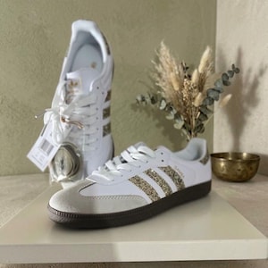 Peut inclure: Baskets blanches avec des rayures et des détails dorés scintillants. Les chaussures ont un bout en daim gris clair et une semelle noire. Une autre basket est visible en arrière-plan, avec une étiquette et des lacets de rechange.
