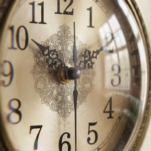 May include: An analog wall clock with a circular brass-toned frame featuring ornate scrollwork details. The clock face has a metallic gold finish with a central decorative floral pattern, black Arabic numerals, and intricate black metal clock hands.