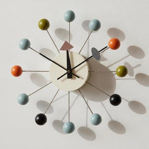 Op de afbeelding: Een wandklok met een crèmekleurige ronde wijzerplaat en zwarte wijzers. Vanuit het midden steken dunne metalen spaken naar buiten, elk voorzien van een bol in lichtblauw, olijfgroen, oranje en zwart, met een roze driehoek als extra accent.
