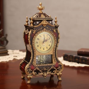 Puede incluir: Un reloj de sobremesa de color rojo oscuro y dorado con un marco arqueado ornamentado y remates decorativos. La esfera presenta números arábigos negros sobre fondo dorado, con un péndulo visible en una ventana de cristal rectangular.