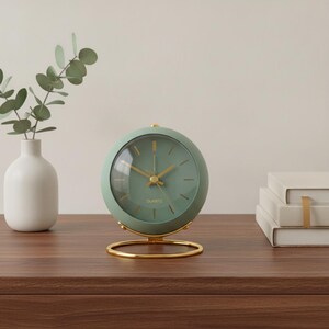 May include: A round sage green analog clock with a gold-toned metal stand and gold hands. The clock face features a clear glass cover, gold hour markers, and the word QUARTZ printed in gold. The clock is positioned on a flat surface.
