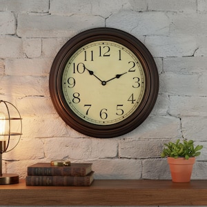 Puede incluir: Un reloj de pared redondo con marco marrón y esfera crema. Los números romanos negros marcan las horas, y las manecillas negras indican la hora. El reloj está montado en una pared de ladrillo blanco. Una pequeña planta en maceta y libros están en una estantería de madera.