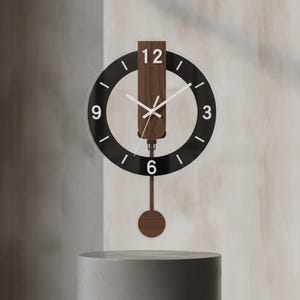 Puede incluir: Un reloj de pared con marco circular negro y números blancos en las posiciones 3, 6, 9 y 12. Un panel vertical de madera oscura atraviesa el centro y sostiene las manecillas blancas. Una varilla fina desciende hasta un péndulo circular de madera.