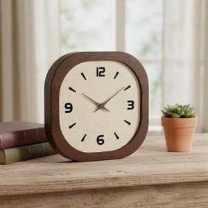 Peut inclure: Une horloge murale carrée aux coins arrondis, dotée d'un cadre en bois brun foncé et d'un cadran couleur crème. L'horloge présente des chiffres noirs aux positions 12, 3, 6 et 9, des graduations noires pour les autres heures et des aiguilles fines.