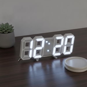 Puede incluir: Un reloj digital blanco con un marco tridimensional y segmentos LED de color blanco brillante que muestran la hora. El reloj cuenta con un panel central con dos pequeños botones circulares y un separador de dos puntos entre los dígitos.
