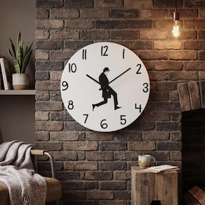 Peut inclure: Une horloge murale ronde blanche avec des chiffres noirs de 1 à 12 et une silhouette noire centrale représentant une personne en costume et chapeau melon en train de marcher. L'horloge possède deux aiguilles noires sur un mur en briques.