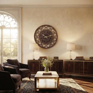 Op de afbeelding: Een grote ronde wandklok met een donkere wijzerplaat en gouden wijzers, voorzien van een ingewikkeld opengewerkt metalen frame. De klok hangt aan een lichte muur boven een donkerhouten dressoir, geflankeerd door twee bijpassende tafellampen.