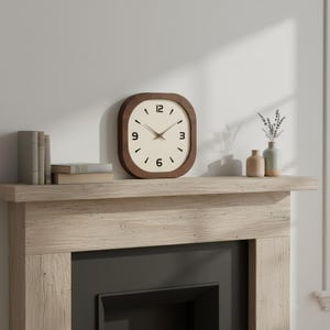 May include: A square wall clock with rounded corners, featuring a dark brown wooden frame, a cream-colored face, and black numerals at the 12, 3, 6, and 9 o'clock positions. The clock has thin, dark-colored hour and minute hands.