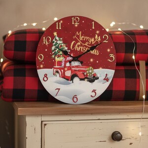 Peut inclure: Une horloge murale ronde à fond rouge représentant un camion rouge vintage transportant un sapin de Noël. Le cadran affiche des chiffres dorés de 1 à 12, des aiguilles noires et le texte doré Merry Christmas. Le motif inclut de la neige blanche.