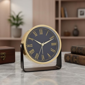 Peut inclure: Une horloge de table ronde dotée d'un cadran bleu marine foncé avec des chiffres romains dorés et des aiguilles dorées assorties. L'horloge est encadrée par un contour en métal doré et repose sur un support en métal noir en forme de U.