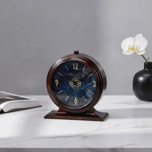 Peut inclure: Une horloge de table ronde avec un cadre en bois brun foncé et un socle. Le cadran présente un motif marbré bleu profond et blanc avec des chiffres dorés aux positions 3, 6, 9 et 12, ainsi que des aiguilles dorées avec un centre décoratif.