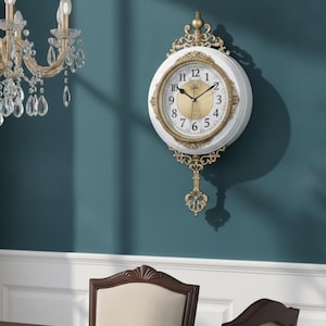 Peut inclure: Une horloge murale ronde avec un cadre blanc et des ornements en filigrane doré en haut et en bas. Le cadran présente un centre doré, une bordure blanche, des chiffres arabes noirs et des aiguilles noires. Un pendule décoratif est suspendu.