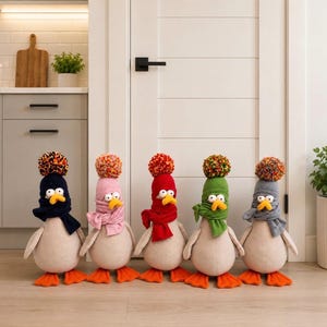 May include: Five plush penguin-like figures standing in a row, each wearing a different coloured knit hat with a matching scarf and a multi-coloured pom-pom on top. The figures have beige bodies, orange beaks, and orange webbed feet.