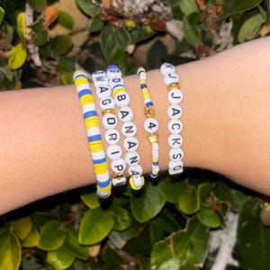 May include: A collection of colorful beaded bracelets. One bracelet features the text "SWAG DRIP", another reads "GO BANANA", and a third says "JACKSO". The bracelets incorporate yellow, blue, and white beads, with gold accents.