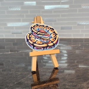 May include: A colorful sticker of a coiled snake, featuring a pattern of yellow, blue, and orange scales. The sticker is displayed on a small wooden easel, set against a gray and white tiled background. The snake's head is visible, with a detailed eye and markings.