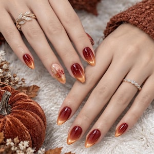 May include: Almond-shaped nails featuring a deep red to orange gradient with a cat-eye effect, one nail accented with a small maple leaf design. A gold-toned leaf-shaped ring with small clear stones and a thin gold band with stones.