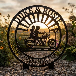 Könnte beinhalten: Ein kreisförmiges schwarzes Metallschild mit der Silhouette von zwei Personen auf einem Motorrad. Der Text DUKE & ROSIE steht oben, und RIDE TOGETHER FORVER EST. 2026 steht unten. Ein Sonnenstrahlen-Design befindet sich hinter dem Motorrad und einem Herzschild.