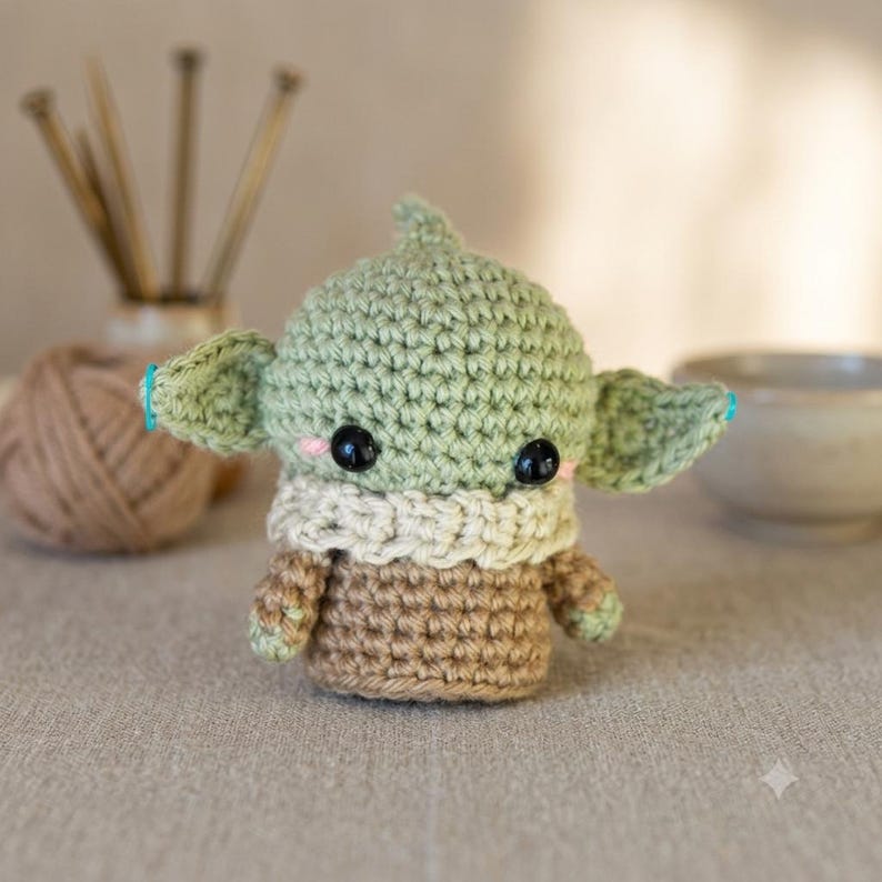 May include: A small, handmade crochet doll featuring a light green head with pointed ears, large black plastic eyes, and pink stitched cheeks. The doll wears a cream-colored textured collar over a brown body, crafted with visible yarn stitches.
