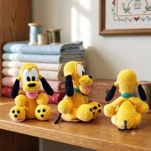 May include: Three crocheted Pluto figures made of yellow yarn with black ears, white muzzle areas, black noses, and pink tongues. Each figure features a teal collar and black paw pads. The figures are displayed in front, side, and back views.