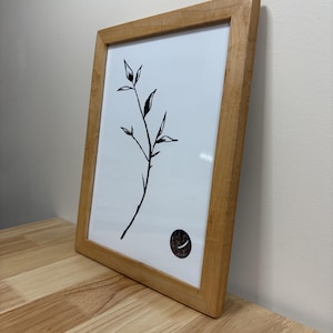 May include: A rectangular wooden frame containing a white print with a black ink illustration of a single plant stem with leaves and a small black circle featuring a white crescent shape positioned in the lower right corner of the artwork.