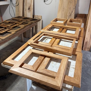 May include: A collection of unfinished light brown wooden picture frames in various rectangular sizes. The frames feature beveled edges and mitered corners, arranged in stacks on a work surface, showcasing the natural grain of the wood.